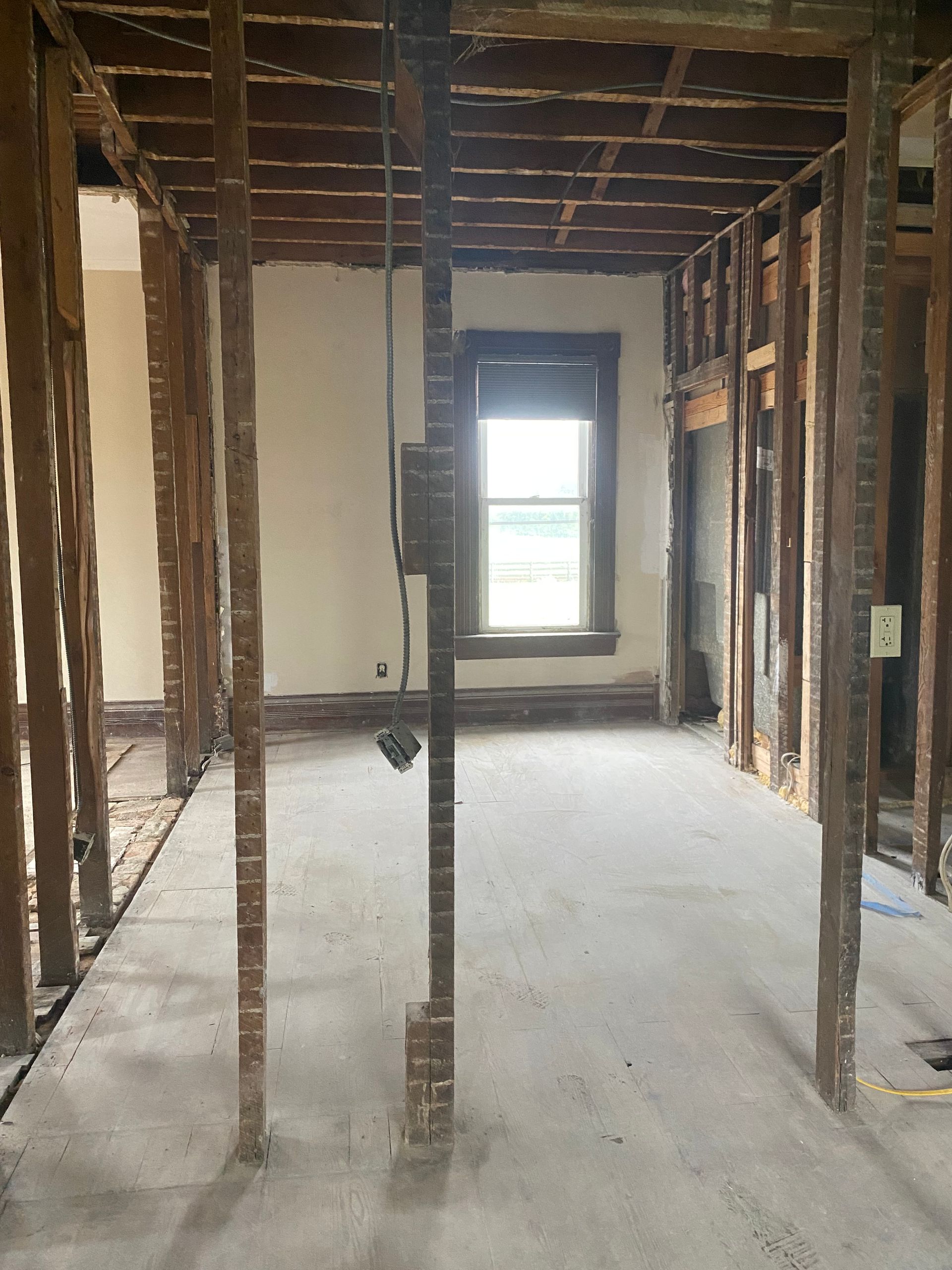 Exposed interior room under renovation with open wall studs, ceiling beams, and a window at the far end.